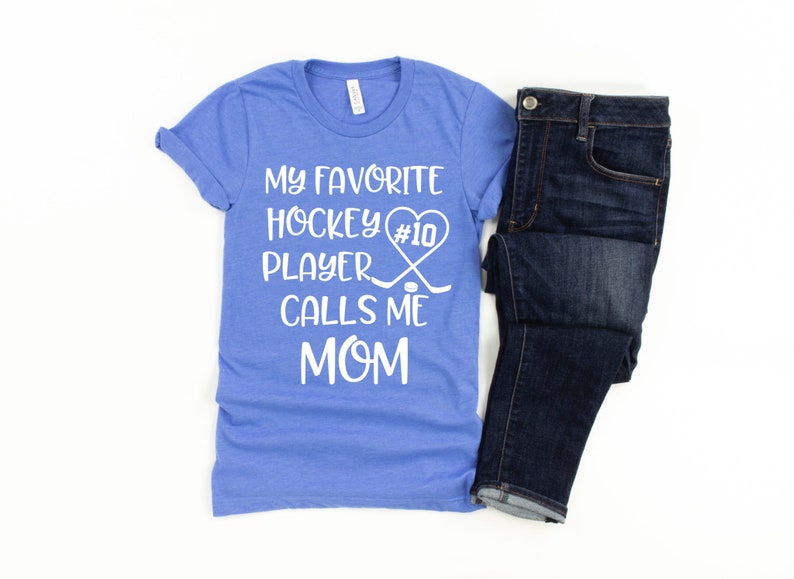 May include: A blue t-shirt with white text that reads "My favorite hockey player #10 calls me mom". The text is in a heart shape with hockey sticks and a puck.