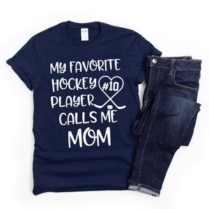 May include: Navy blue t-shirt with white text that reads "My favorite hockey player #10 calls me mom". The text is accompanied by a heart with a hockey stick and puck graphic.