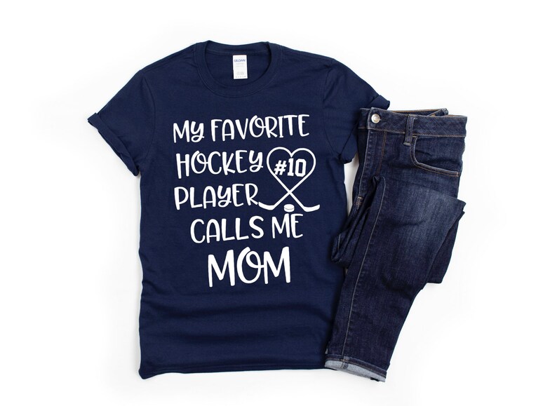May include: Navy blue t-shirt with white text that reads "My favorite hockey player #10 calls me mom". The text is accompanied by a heart with a hockey stick and puck graphic.