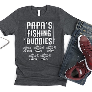 Puede incluir: Una camiseta gris oscuro con el texto "Papa's Fishing Buddies" y los nombres "Carter, Jayce, Cody, Harper, Tracy" impresos en ella. Cada nombre va acompa&ntilde;ado de una peque&ntilde;a ilustraci&oacute;n de un pez.