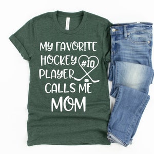 May include: A green t-shirt with white text that reads "My favorite hockey player #10 calls me mom". The text is surrounded by a heart and hockey sticks. A pair of blue jeans is folded on the right side of the image.