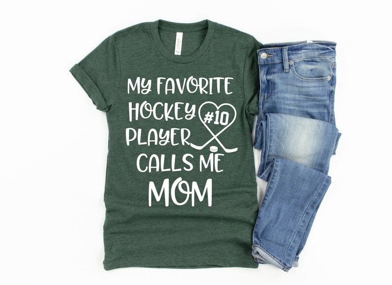 May include: A green t-shirt with white text that reads "My favorite hockey player #10 calls me mom". The text is surrounded by a heart and hockey sticks. A pair of blue jeans is folded on the right side of the image.