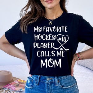 May include: Black t-shirt with white text that reads "My favorite hockey player #10 calls me mom". The text is surrounded by a heart with hockey sticks and a puck.