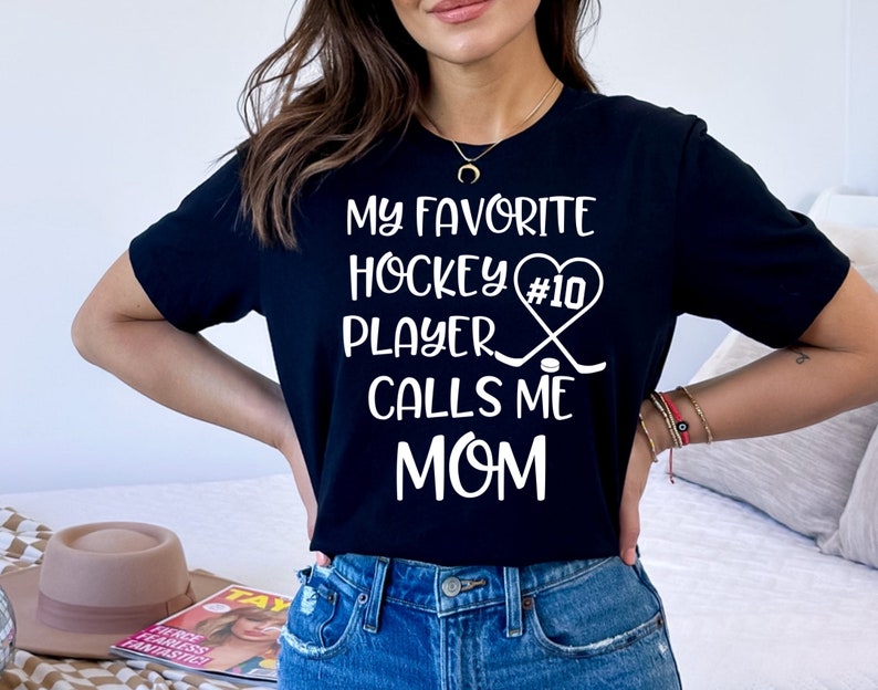 May include: Black t-shirt with white text that reads "My favorite hockey player #10 calls me mom". The text is surrounded by a heart with hockey sticks and a puck.