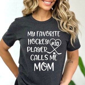 May include: A dark gray t-shirt with white text that reads "My favorite hockey player #10 calls me mom". The text is accompanied by a graphic of a hockey stick and puck.