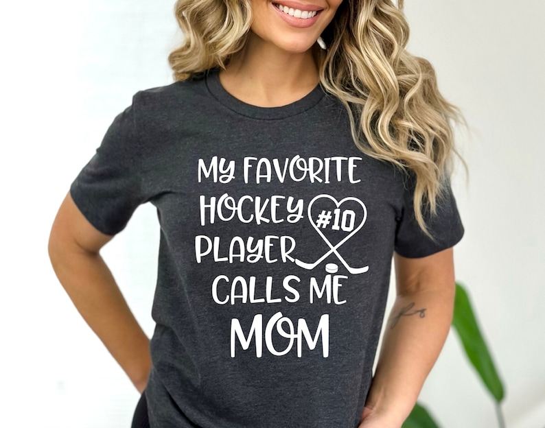 May include: A dark gray t-shirt with white text that reads "My favorite hockey player #10 calls me mom". The text is accompanied by a graphic of a hockey stick and puck.