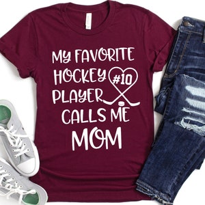 May include: Burgundy t-shirt with white text that reads "My favorite hockey player #10 calls me mom". The text is accompanied by a heart, hockey sticks, and a puck.
