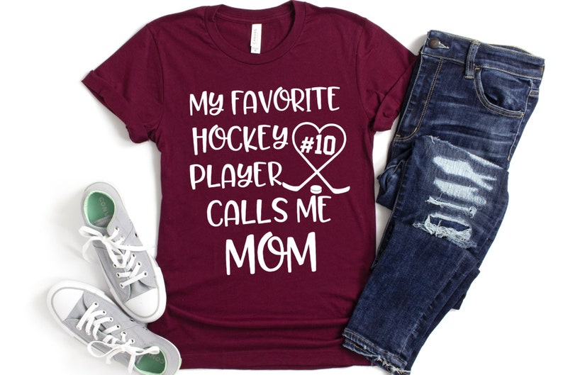 May include: Burgundy t-shirt with white text that reads "My favorite hockey player #10 calls me mom". The text is accompanied by a heart, hockey sticks, and a puck.