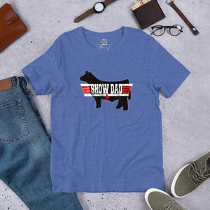 May include: A blue t-shirt with a black silhouette of a cow with the text "SHOW DAD" in white letters above the cow. The cow is in a red, white, and blue graphic that resembles a fighter jet.