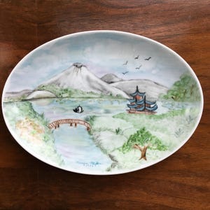May include: An oval ceramic dish featuring a hand-painted landscape scene with a large mountain, a blue lake, a wooden bridge, a small boat, and a pagoda. The artwork includes green trees, soft clouds, and birds in the sky. The signature reads Morgan 1981.