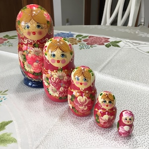 May include: A set of five Russian nesting dolls, or Matryoshka dolls, arranged in a row. The dolls are painted with floral designs in shades of red, pink, and white, with gold accents. Each doll features a painted face.