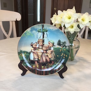 May include: A round, colorful ceramic plate with a depiction of four children in traditional German clothing dancing around a maypole. The plate is on a wooden stand and has a gold rim.