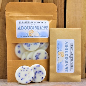 Peut inclure: Deux sachets en papier kraft avec des étiquettes indiquant "12 Pastilles Parfumées Adoucissant" et "2 Pastilles Parfumées Adoucissant". Les sachets contiennent des pastilles de cire blanche avec des points bleus.