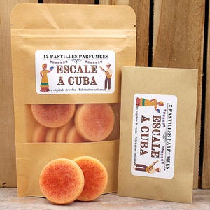 Peut inclure: Deux emballages en papier brun et un sac en plastique transparent remplis de pastilles rondes de couleur orange. Les emballages portent des étiquettes avec le texte "12 PASTILLES PARFUMÉES ESCALE À CUBA". L'arrière-plan est une surface en bois.