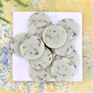 May include: A pile of round, light green soap bars with dried lavender flowers and herbs embedded in them. The soap bars are arranged on a white marble surface.