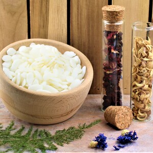 May include: A wooden bowl filled with white wax flakes, two glass bottles with cork stoppers, one filled with dried red flower petals, the other with dried yellow flower petals, and a green fern on a wooden surface.