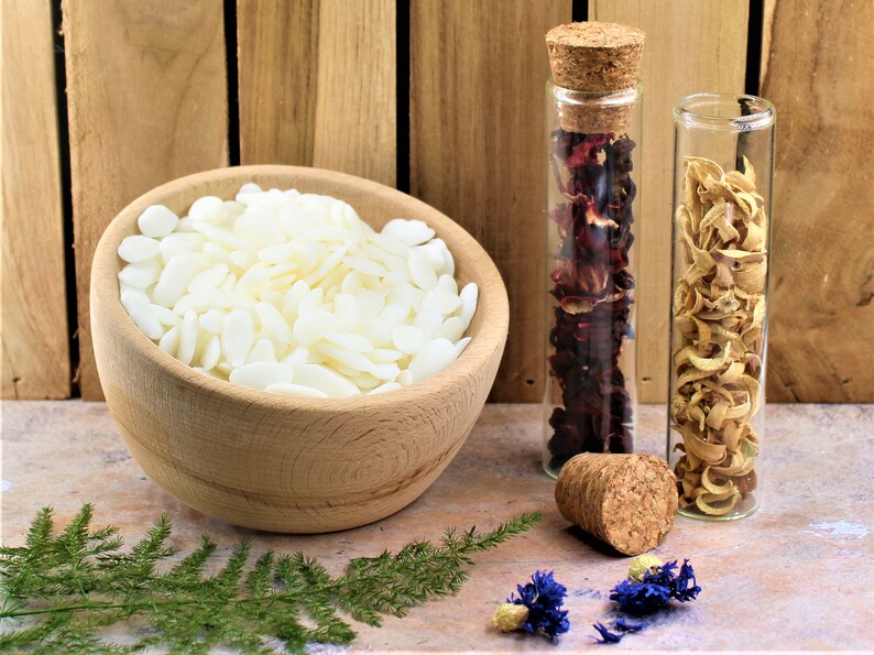 May include: A wooden bowl filled with white wax flakes, two glass bottles with cork stoppers, one filled with dried red flower petals, the other with dried yellow flower petals, and a green fern on a wooden surface.