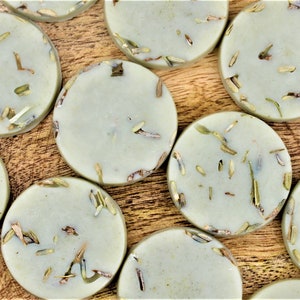 May include: A close-up of a group of round, light green soap bars with dried herbs embedded in them. The soap bars are arranged on a wooden surface.