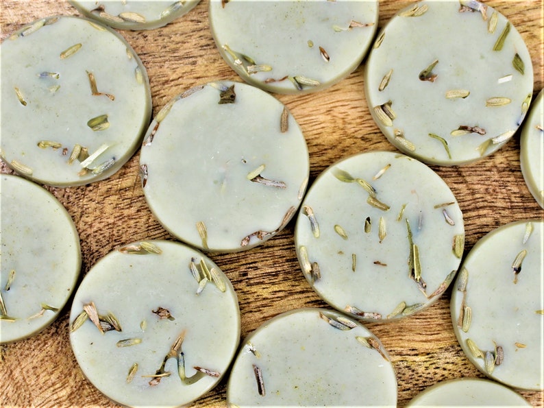 May include: A close-up of a group of round, light green soap bars with dried herbs embedded in them. The soap bars are arranged on a wooden surface.