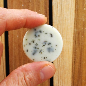May include: A round, white soap bar with green flecks. The soap is being held by a hand.