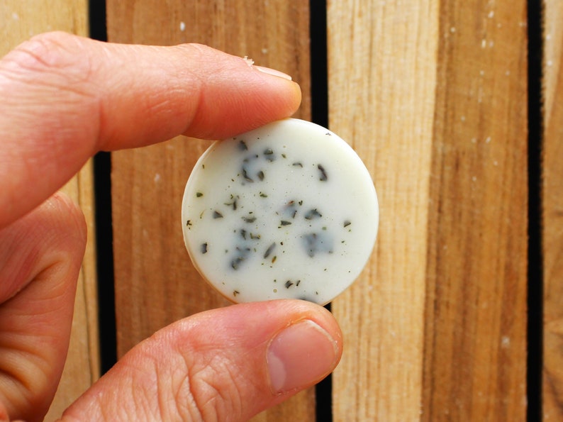 May include: A round, white soap bar with green flecks. The soap is being held by a hand.