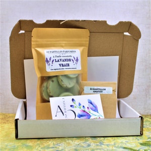 May include: A white cardboard box with a brown paper bag inside. The bag contains 12 scented wax melts with a lavender scent. The bag has a label that reads "12 Pastilles Parfum&eacute;es &agrave; l'huile essentielle Lavande Vraie. Cire v&eacute;g&eacute;tale de soja - Fabrication artisanale". The box also contains a small brown paper envelope with a label that reads "&Eacute;chantillon Gratuit".