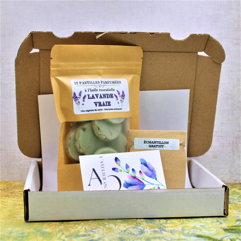 May include: A white cardboard box with a brown paper bag inside. The bag contains 12 scented wax melts with a lavender scent. The bag has a label that reads "12 Pastilles Parfum&eacute;es &agrave; l'huile essentielle Lavande Vraie. Cire v&eacute;g&eacute;tale de soja - Fabrication artisanale". The box also contains a small brown paper envelope with a label that reads "&Eacute;chantillon Gratuit".