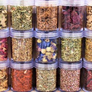 May include: Twenty clear glass jars filled with various dried herbs and flowers. The jars are arranged in three rows of six jars each. The herbs and flowers are a variety of colours, including red, pink, yellow, green, and brown.