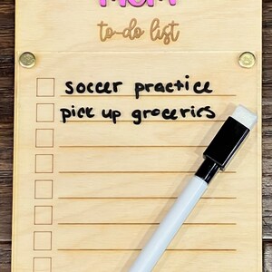 Pode incluir: Um quadro de madeira para lista de tarefas com a palavra "MOM" em letras cor-de-rosa e o texto "to-do list" escrito em castanho. O quadro tem um marcador de apagamento a seco branco e uma borracha preta. O quadro tem uma lista de tarefas escritas nele, incluindo "soccer practice" e "pick up groceries".