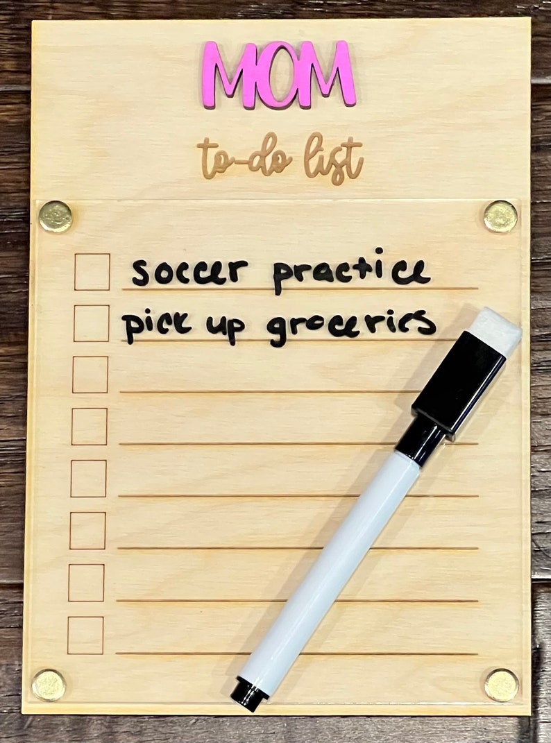Pode incluir: Um quadro de madeira para lista de tarefas com a palavra "MOM" em letras cor-de-rosa e o texto "to-do list" escrito em castanho. O quadro tem um marcador de apagamento a seco branco e uma borracha preta. O quadro tem uma lista de tarefas escritas nele, incluindo "soccer practice" e "pick up groceries".