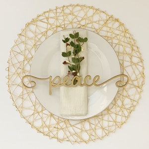 May include: A gold wire placemat with a white plate and a white napkin. A sprig of greenery and a wooden sign with the word "peace" are on the plate.