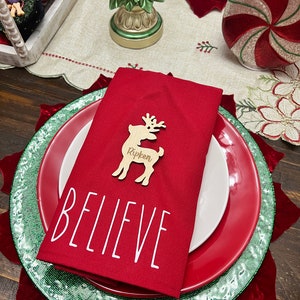 May include: A red napkin with the word "BELIEVE" printed on it. A wooden reindeer place card with the name "Rippen" is on top of the napkin. The napkin is on top of a green and red plate.