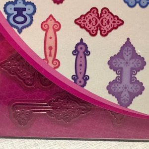 May include: A set of metal cutting dies for crafting, featuring intricate shapes including ornate keyhole plates, decorative hinges, and a skeleton key. The dies are presented on a white card with a bright pink diagonal accent and a textured base.
