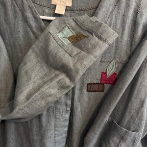 May include: A light gray long-sleeved shirt featuring a V-neckline and decorative stitching. The garment is adorned with several small, colorful fabric patches in shades of blue, pink, and brown, each embellished with a stitched leaf motif.