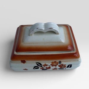 Peut inclure: Une boîte à bijoux carrée en céramique avec un couvercle. La boîte est principalement de couleur crème avec une bordure marron et une poignée blanche sur le couvercle. Des motifs floraux orange et noirs décorent la base de la boîte.