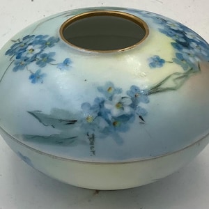 Op de afbeelding: Een ovaalvormige keramische container met een deksel, beschilderd met lichtblauwe en witte tinten. Delicate blauwe bloempatronen sieren het oppervlak en een gouden rand omcirkelt de opening. De container heeft een zachte, pastelachtige uitstraling.