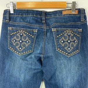 May include: Close-up of the back of blue denim jeans. The jeans have decorative stitching and embellishments on the back pockets. The pockets feature a floral design with small, round studs and rhinestones. The brand tag "South" is visible.