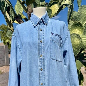 May include: Light blue denim shirt with long sleeves, a button-down front, and a collared neckline. The shirt features a chest pocket on the left side and a small logo with text. The shirt is displayed on a mannequin.