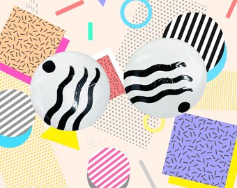 May include: Two white round earrings with black wave and dot designs. The earrings are set against a background of geometric shapes in various colors, including pink, yellow, blue, and gray. The overall aesthetic is reminiscent of the 1980s.