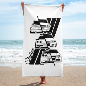 May include: White beach towel featuring a black and white graphic of three Skyline cars. The word "Skyline" is written in a stylized font. Black diagonal stripes are behind the cars.
