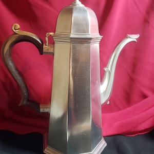 May include: A tall, silver-colored coffee pot with a wooden handle and a decorative spout. The pot has a geometric design and a domed lid with a finial. The handle is dark brown and curved. The pot is likely made of metal.