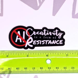May include: A black and white sticker with the text "AI" crossed out in a red circle, and the words "Creativity is a form of RESISTANCE". A green ruler with inch markings is below the sticker.