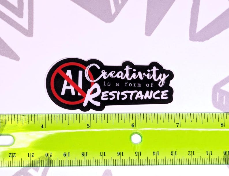 May include: A black and white sticker with the text "AI" crossed out in a red circle, and the words "Creativity is a form of RESISTANCE". A green ruler with inch markings is below the sticker.