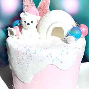 May include: A festive cake with a pink base and white frosting, decorated with a polar bear wearing a pink hat, an igloo, and pink and blue ornaments. Two pink bottle brush trees stand behind the bear and igloo.