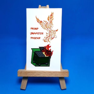 May include: A white card on a wooden easel features a graphic of a phoenix and a dumpster with flames. The text reads "Proud Dumpster Phoenix" in red. The phoenix is orange and red.