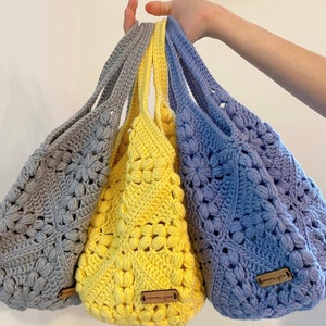 May include: Three crocheted tote bags in gray, yellow, and purple with a flower pattern. Each bag has a brown leather tag with the text "handmade" on it.