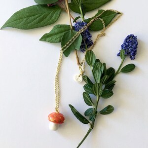 May include: A gold chain necklace with a red and white mushroom pendant. The necklace is laying on a white surface with green leaves and blue flowers.