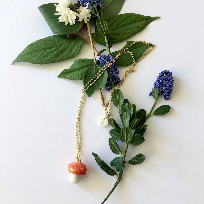 May include: A gold chain necklace with a red and white mushroom pendant. The necklace is laying on a white surface with green leaves and blue flowers.