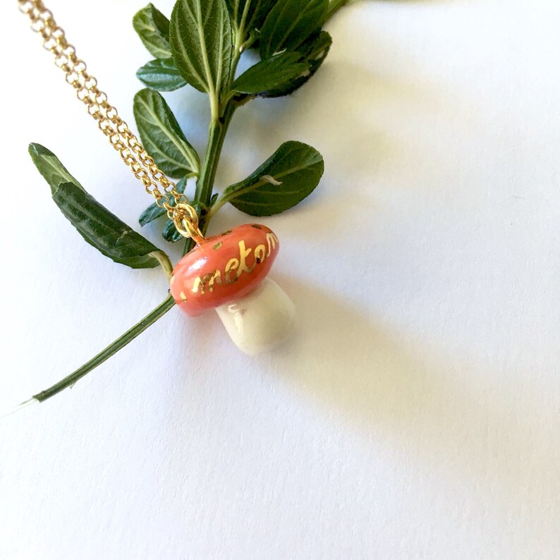 May include: A gold chain necklace with a red and white ceramic mushroom pendant. The mushroom has gold lettering that says "meton".