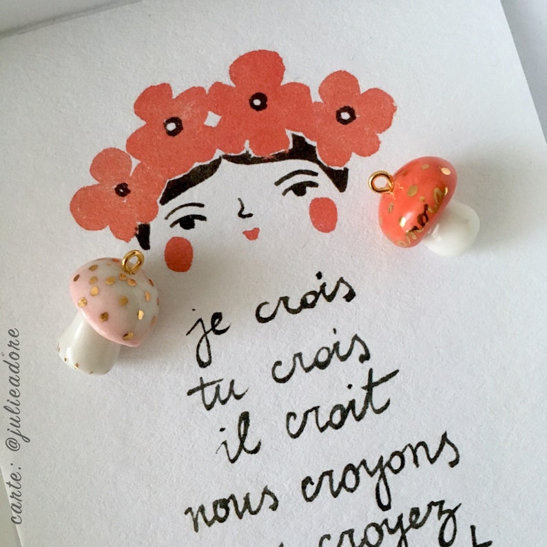 May include: Two ceramic mushroom charms with gold accents. One mushroom is pink with gold dots and the other is red with gold speckles. The charms are on a white background with a hand-drawn illustration of a woman with a flower crown and the text "je crois tu crois il croit nous croyons croyez".